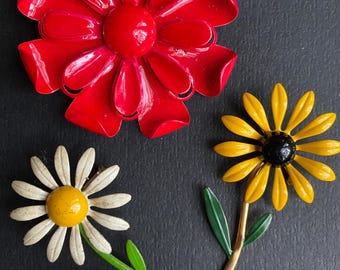 May include: Three flower-shaped brooches. A large, bright red flower brooch with rounded petals. A daisy brooch with white petals and a yellow center. A yellow flower brooch with a black center and green leaves.