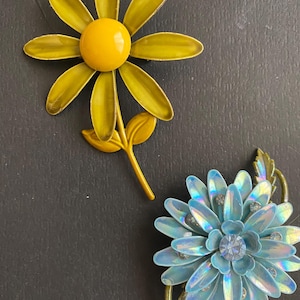 May include: Two flower-shaped brooches. One is yellow with a yellow center and a stem with leaves. The other is blue with iridescent petals and a central rhinestone. Both are set against a dark background.