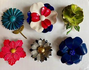 May include: Six vintage metal flower brooches in various colors, including blue, green, red, white, and gray. The brooches are all different shapes and sizes, and some have a raised center.