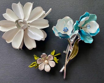 May include: Three floral brooches in a variety of designs. One is a large white daisy, another features two blue and white flowers with blue crystal centers, and the third is a small pink flower with a pearl center and green leaves.