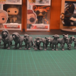 Può includere: Una collezione di nove figurine di leone grigie, ognuna con una posa ed un'espressione uniche. Le figurine sono disposte in fila su una superficie verde.