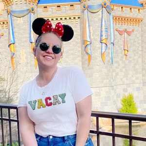 May include: A woman wearing a white t-shirt with the word "VACAY" in colorful letters, blue denim shorts, and a black fanny pack. She is wearing a red Minnie Mouse headband with white polka dots and sunglasses. She is standing in front of a large castle with a moat.