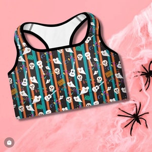 May include: A black sports bra with a colorful pattern of skulls, guitars, ghosts, and fireworks on a striped background. The bra has a racerback style and is made of a stretchy, breathable fabric.