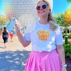 Puede incluir: Una mujer con una camiseta blanca con un diseño de Mickey Mouse de calabaza de purpurina dorada y una falda plisada rosa. Está de pie frente a una gran esfera blanca con un patrón de arcoíris.