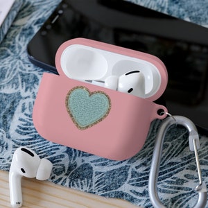 Puede incluir: Una funda de silicona rosa para auriculares inalámbricos con un diseño de corazón turquesa brillante. La funda tiene un mosquetón plateado.