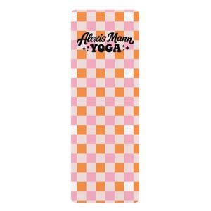 May include: A rectangular yoga mat featuring a repeating checkered pattern in shades of orange, light pink, and white. The text Alexis Mann YOGA is printed in black in the upper centre, flanked by small four-pointed star icons on each side.
