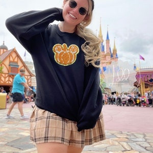 Pode incluir: Uma camisa de frio preta com um design de cabeça de Mickey Mouse em forma de abóbora em glitter laranja e dourado. A camisa de frio está sendo usada por uma pessoa com cabelo loiro e óculos de sol, em pé na frente de um castelo.