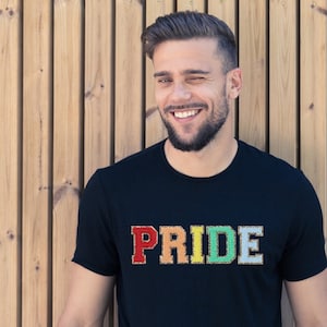 Op de afbeelding: Een zwart T-shirt met het woord "PRIDE" erop geschreven in regenboogkleuren. De letters zijn omrand met goud.