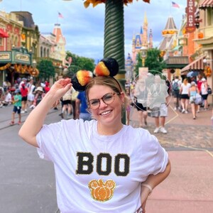 Puede incluir: Una camiseta blanca con la palabra "BOO" en purpurina negra y una cabeza de Mickey Mouse de calabaza en purpurina naranja y dorada. La persona que lleva la camiseta está en un parque de atracciones con decoraciones de otoño.