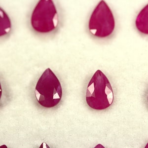 May include: Ten pear-shaped ruby gemstones, each with a deep red hue, are arranged in two rows on a white surface.