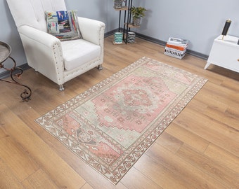 Puede incluir: Una alfombra rectangular de estilo vintage con un diseño de medallón rosa central y un borde beige. La alfombra está colocada sobre un suelo de madera, con un sillón blanco y una mesa auxiliar en el fondo. La alfombra mide unos 90 x 150 cm.