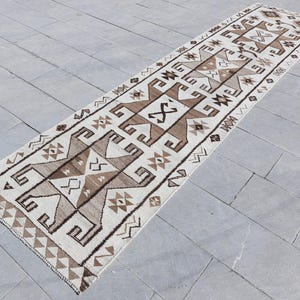 Könnte beinhalten: Ein weißer und brauner Läufer mit geometrischen Mustern. Der Teppich hat einen traditionellen Stil und zeigt ein sich wiederholendes Muster aus Dreiecken, Quadraten und Rauten.