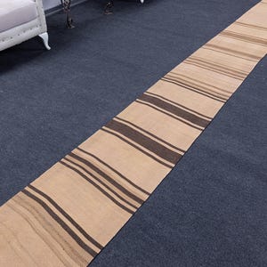 May include: A long, narrow runner rug with brown and beige stripes. The rug is made of a woven material and has a fringed edge.
