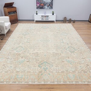 May include: A large, beige rug with a light blue and brown geometric pattern. The rug has a fringe border and is laid out on a wooden floor.