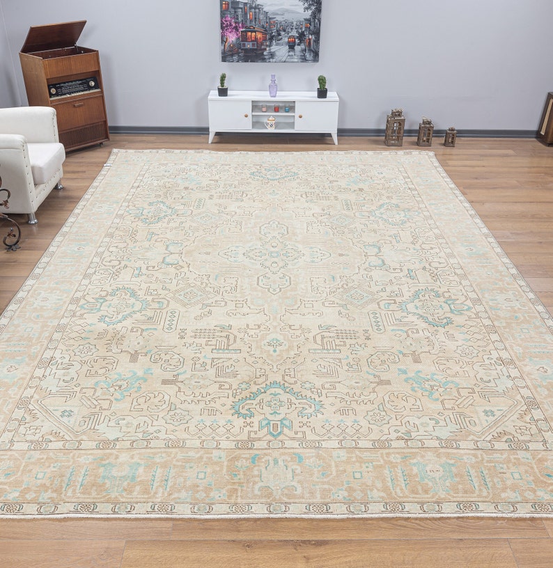 May include: A large, beige rug with a light blue and brown geometric pattern. The rug has a fringe border and is laid out on a wooden floor.
