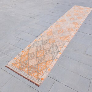 May include: A long, rectangular rug with a diamond pattern in shades of orange and gray. The rug is laid out on a gray tiled floor.