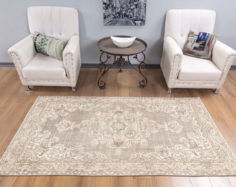 May include: A beige and brown patterned rug with a floral design. The rug is placed on a hardwood floor in front of two white armchairs and a metal table.