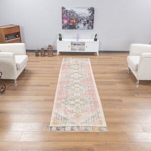 Op de afbeelding: Een witloperkleed met een geometrisch patroon in roze, grijs en geel. Het kleed ligt op een lichtbruine houten vloer in een woonkamer met twee witte fauteuils en een wit tv-meubel.