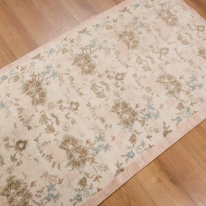 May include: Rectangular rug with a floral pattern in cream, brown, and blue hues. The rug has a pink border and is placed on a wooden floor. The floral design is spread across the rug's surface.