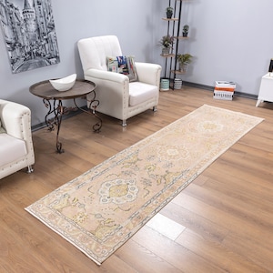 May include: A light beige runner rug with a floral pattern in shades of cream, yellow, and brown. The rug is laid out on a hardwood floor in a living room setting.