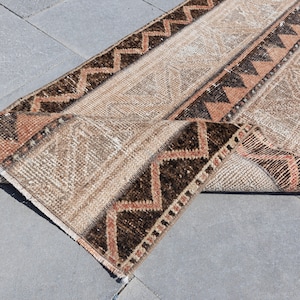 Può includere: Un tappeto da corridoio beige e marrone con un motivo geometrico. Il tappeto è piegato a metà e ha un bordo con frange.
