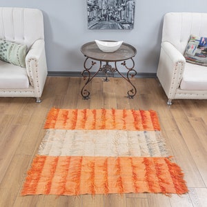 May include: A bright orange and cream striped rug with fringe edges. The rug is placed in front of two white armchairs and a round metal table with a white bowl on top.
