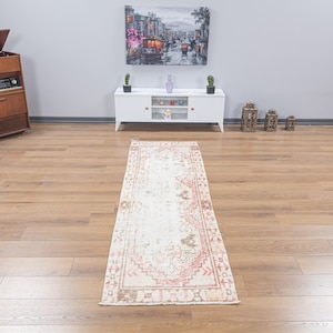 May include: A white and pink vintage runner rug with a geometric pattern. The rug is laid out on a wooden floor in a room with a white entertainment unit and a painting on the wall.