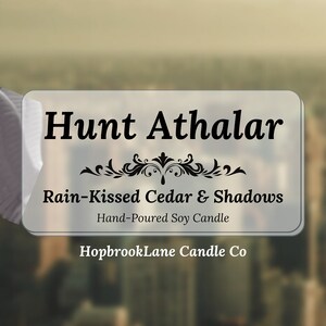 Op de afbeelding: Een wit label met zwarte tekst die "Hunt Athalar", "Rain-Kissed Cedar & Shadows", "Hand-Poured Soy Candle" en "HopbrookLane Candle Co" zegt.