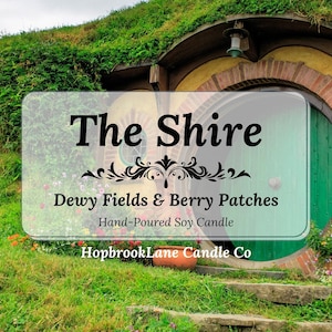 Op de afbeelding: Een handgegoten sojawas-kaars met een groen label waarop staat "The Shire Dewy Fields & Berry Patches Hand-Poured Soy Candle Hopbrook Lane Candle Co.". De kaars staat voor een hobbitwoning met een ronde groene deur.