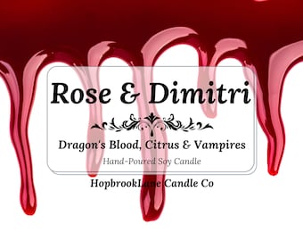 May include: A white background with a decorative border of dripping red liquid. The text "Rose & Dimitri" is displayed above "Dragon's Blood, Citrus & Vampires" and "Hand-Poured Soy Candle". The brand name "Hopbrook Lane Candle Co" is at the bottom.