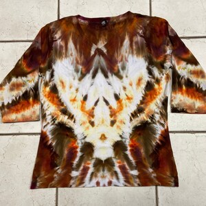 May include: A V-neck, three-quarter sleeve shirt with a tie-dye pattern. The shirt features a blend of colors, including white, brown, orange, and burgundy. The tie-dye design is symmetrical, with a central white area and radiating patterns.
