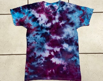 May include: A short-sleeved t-shirt with a tie-dye pattern in shades of blue, purple, and white. The shirt has a crew neck and a classic fit. The tie-dye design creates a unique, swirling effect across the fabric.