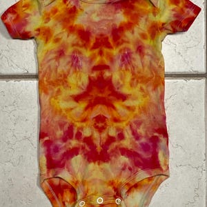 May include: A short-sleeved baby bodysuit with a vibrant tie-dye pattern in shades of red, orange, and yellow. The bodysuit has a round neckline and three snap closures at the bottom. The fabric appears to be soft and comfortable.