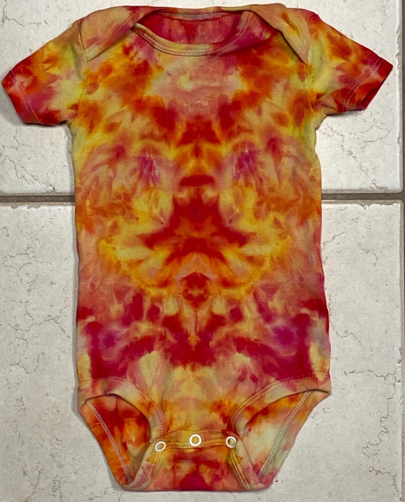 May include: A short-sleeved baby bodysuit with a vibrant tie-dye pattern in shades of red, orange, and yellow. The bodysuit has a round neckline and three snap closures at the bottom. The fabric appears to be soft and comfortable.