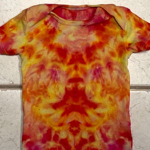 May include: A tie-dye baby onesie in vibrant shades of red, orange, yellow, and pink. The short-sleeved garment features a round neckline and a symmetrical pattern. The fabric appears soft and comfortable.