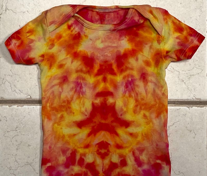 May include: A tie-dye baby onesie in vibrant shades of red, orange, yellow, and pink. The short-sleeved garment features a round neckline and a symmetrical pattern. The fabric appears soft and comfortable.