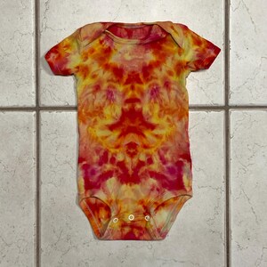 May include: A tie-dye baby bodysuit in shades of red, orange, and yellow. The short-sleeved onesie has three snap closures at the bottom. The fabric features a symmetrical pattern, with the colors blending together.