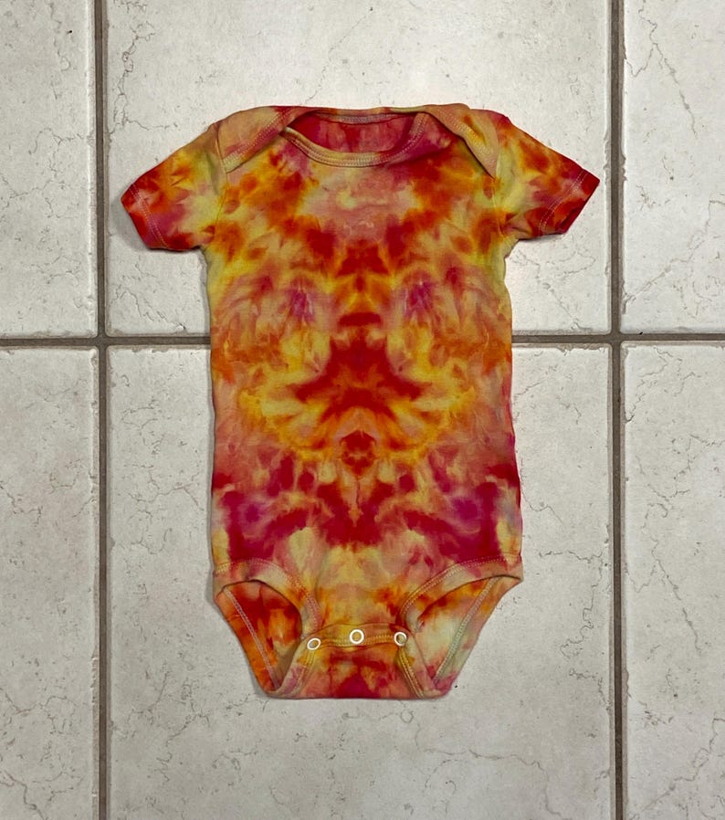 May include: A tie-dye baby bodysuit in shades of red, orange, and yellow. The short-sleeved onesie has three snap closures at the bottom. The fabric features a symmetrical pattern, with the colors blending together.