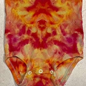 May include: A tie-dye baby bodysuit in shades of red, orange, and yellow. The one-piece garment features a round neckline, short sleeves, and three snap closures at the bottom. The fabric has a vibrant, swirling pattern.