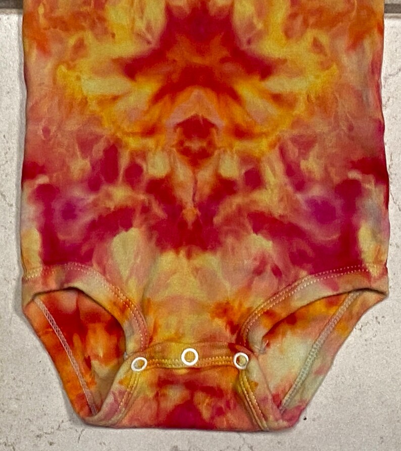 May include: A tie-dye baby bodysuit in shades of red, orange, and yellow. The one-piece garment features a round neckline, short sleeves, and three snap closures at the bottom. The fabric has a vibrant, swirling pattern.