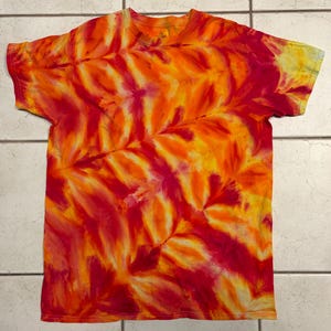 May include: A short-sleeved t-shirt with a vibrant tie-dye pattern in shades of red, orange, and yellow. The design features a leaf-like pattern, creating a dynamic and colorful visual effect. The shirt is made of a soft material.