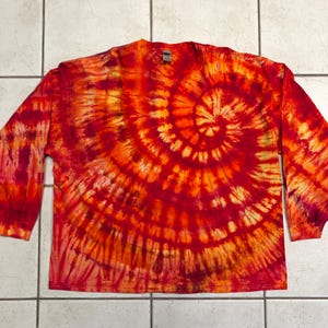 May include: A long-sleeved shirt with a vibrant tie-dye pattern in shades of red, orange, and yellow. The spiral design radiates from the center, creating a bold and eye-catching visual effect. The shirt is laid flat, showcasing its full design.