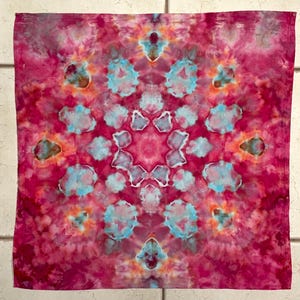 May include: A square, tie-dye textile with a symmetrical pattern. The color palette includes shades of pink, red, and blue. The design features a central star-like shape surrounded by floral-like motifs. The fabric has a soft, textured appearance.
