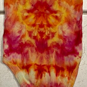May include: A tie-dye baby onesie in vibrant shades of red, orange, and yellow. The fabric features a symmetrical pattern with a blend of warm colors. The onesie has a round neckline and short sleeves.