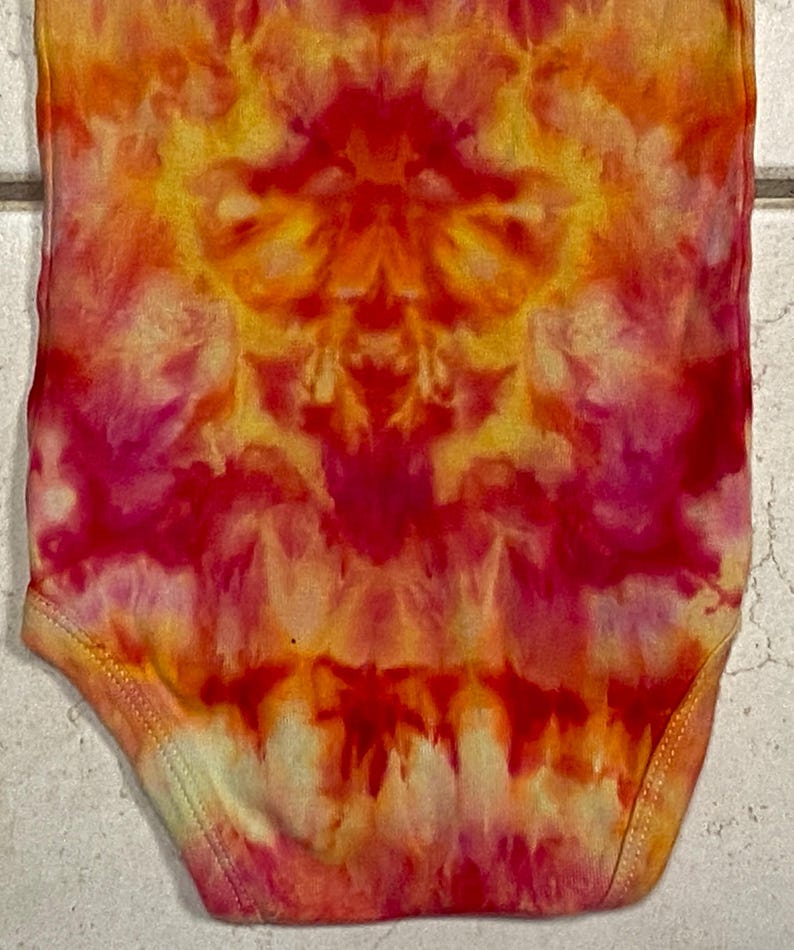 May include: A tie-dye baby onesie in vibrant shades of red, orange, and yellow. The fabric features a symmetrical pattern with a blend of warm colors. The onesie has a round neckline and short sleeves.