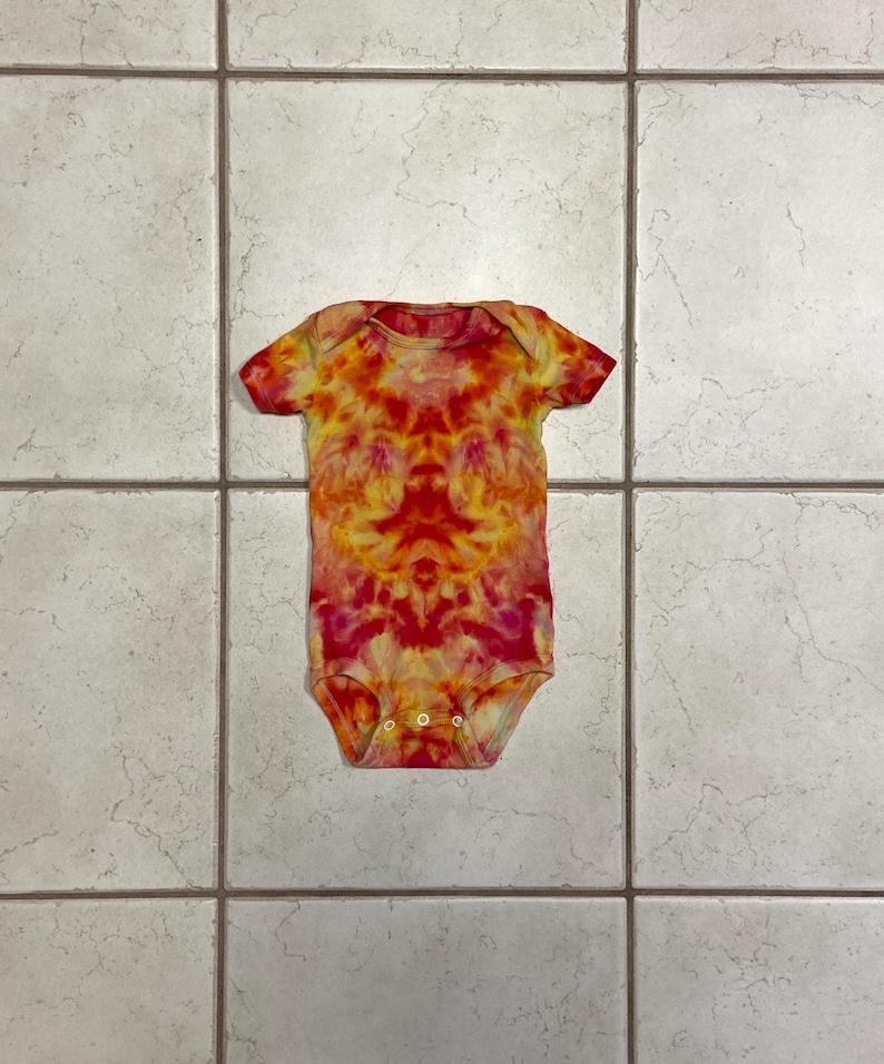 May include: A tie-dye baby onesie in shades of red, orange, and yellow. The short-sleeved garment has a vibrant, swirled pattern. The onesie is buttoned at the bottom and is laying flat on a tiled surface.