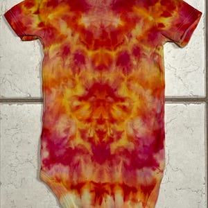 May include: A tie-dye baby onesie in shades of red, orange, and yellow. The short-sleeved garment features a symmetrical pattern, with the colors blending and swirling together. The onesie is made of soft fabric.