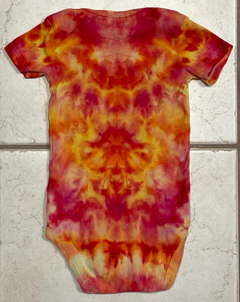 May include: A tie-dye baby onesie in shades of red, orange, and yellow. The short-sleeved garment features a symmetrical pattern, with the colors blending and swirling together. The onesie is made of soft fabric.