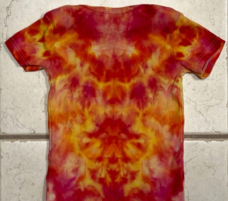 May include: A short-sleeved t-shirt with a tie-dye pattern in shades of red, orange, and yellow. The shirt has a crew neck and a symmetrical design. The fabric appears to be soft and lightweight, suitable for casual wear.
