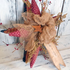 May include: A red, white, and blue star-shaped fabric decoration with a burlap bow and sprigs of dried flowers. The star is made of multiple layers of fabric and has a rustic, vintage look.